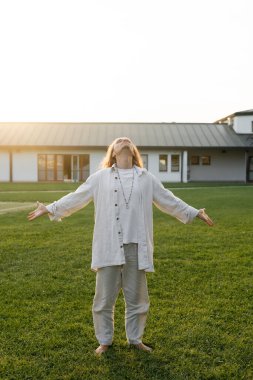 full length of man in linen clothes meditating with outstretched hands and raised head on green lawn near countryside cottage clipart