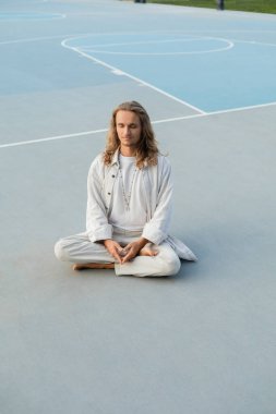 Uzun saçlı, uzun boylu, şık bir adam. Açık stadyumda yoga pozisyonunda meditasyon yapıyor.