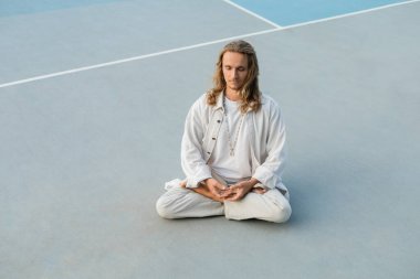 Beyaz pamuklu elbiseli uzun saçlı adam dışarıda Lotus pozu vererek yoga yapıyor.