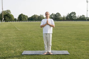 full length of barefoot man in linen pants meditating in mountain pose with praying hands on yoga mat on green lawn clipart