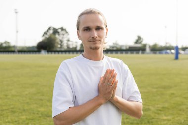 young and positive man in white t-shirt showing anjali mudra gesture and looking at camera on blurred outdoor stadium clipart