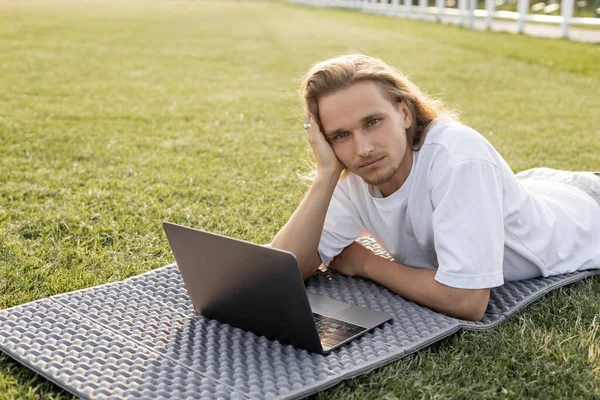 stock image dreamy yoga man looking at camera while lying near laptop on green grass outdoors