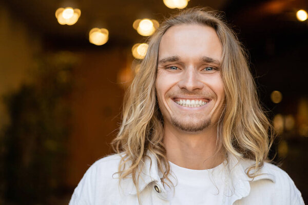 portrait of overjoyed long haired yoga man smiling at camera near blurred lights outdoors
