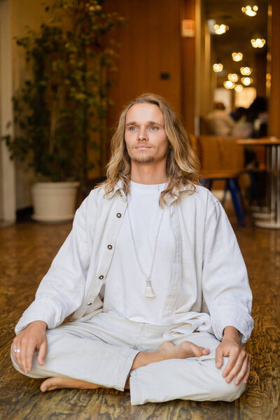 молодой и стильный мужчина с длинными светлыми волосами, глядя в сторону во время медитации в легкой позе в коттедже с размытыми огнями