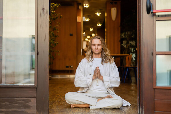 young long haired man meditating in easy pose with anjali mudra gesture at entrance of house