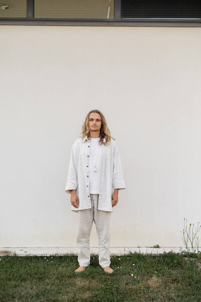 full length of barefoot and fair haired man in linen clothes standing near white wall outdoors