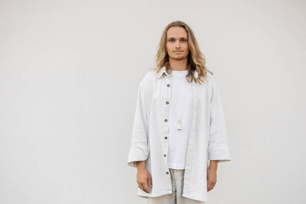 stylish yoga man with long fair hair standing near white wall and looking at camera