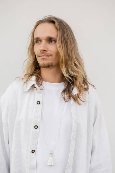 portrait of young long haired yoga man in linen shirt looking away near white wall outdoors