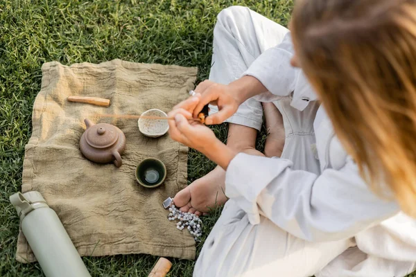 top view of yoga man holding scented stick near linen rug with clay teapot and bowls on grassy lawn