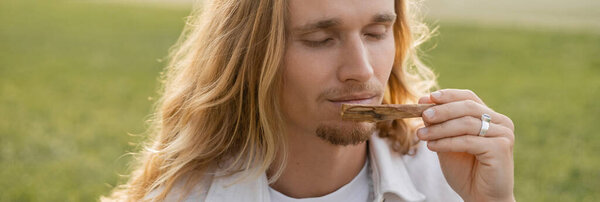 young man with long hair and closed eyes smelling fragrant palo santo stick outdoors, banner