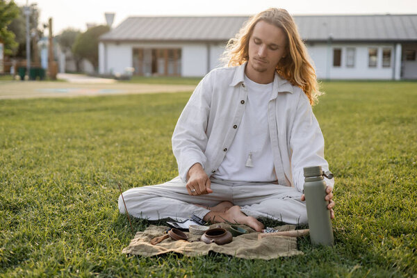 long haired man in white linen clothes holding thermos near ceramic teapot and bowls during tea ceremony in countryside
