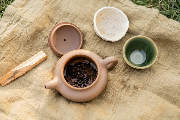 top view of clay bowls and teapot with puer tea near palo santo stick on linen rug outdoors