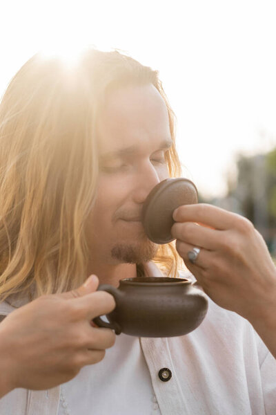 pleased long haired man with closed eyes holding oriental teapot and enjoying flavor of puer tea in sunshine outdoors