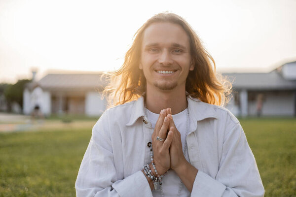carefree long haired man with mala beads showing anjali mudra gesture and smiling at camera outdoors