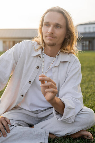 длинноволосый мужчина в льняной одежде трогает бусины и смотрит в сторону во время медитации на открытом воздухе