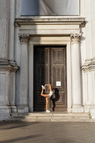 Side view of sportswoman in bodysuit and sunglasses stretching near building in Venice — Stock Photo