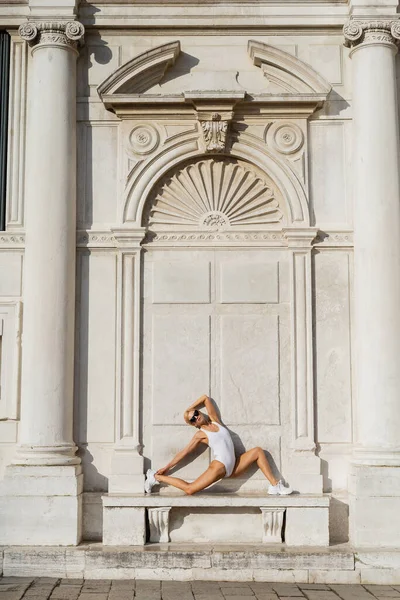 Blonde sportswoman in sunglasses and bodysuit stretching near building on street in Venice — Stock Photo