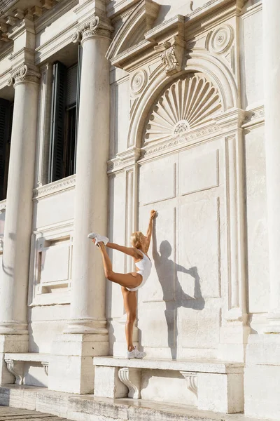 Вид сбоку на стройную блондинку в белом халате и бодибилдере, растягивающуюся возле здания в Италии — стоковое фото