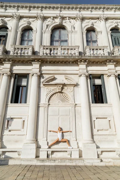 Vista lateral de la deportista en traje de cuerpo y gafas de sol de entrenamiento en el banco cerca de la construcción en Venecia - foto de stock