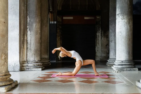Mulher loira em bodysuit branco praticando ioga no tapete perto do edifício na Itália — Fotografia de Stock