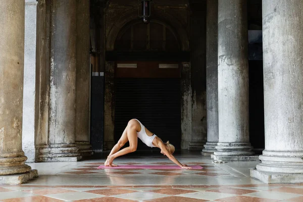 Femme blonde s'étirant tout en pratiquant le yoga en body blanc sur le tapis autour du bâtiment avec des colonnes à Venise — Photo de stock
