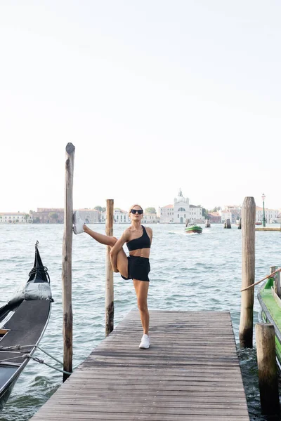 Flexible Sportlerin mit Sonnenbrille, weißen Turnschuhen, schwarzem bauchfreien Oberteil und kurzen Hosen beim Training auf einem Pier in Venedig — Stockfoto