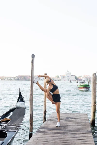 Blonde sportswoman in sunglasses, white sneakers, black crop top and shorts training on pier in Venice — Stock Photo