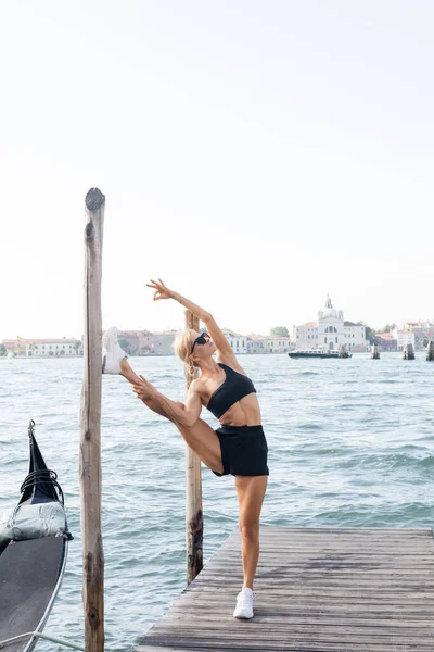 Fit sportswoman in sunglasses, white sneakers, black crop top and shorts stretching on pier in Venice — Stock Photo
