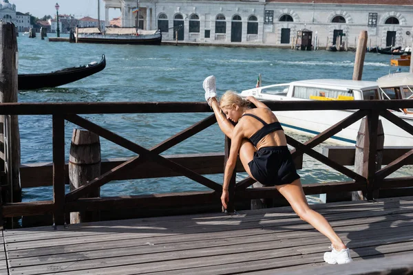 Seitenansicht einer blonden Sportlerin in weißen Turnschuhen, schwarzem bauchfreiem Oberteil und Shorts, die sich auf einem Pier in Italien ausdehnt — Stockfoto