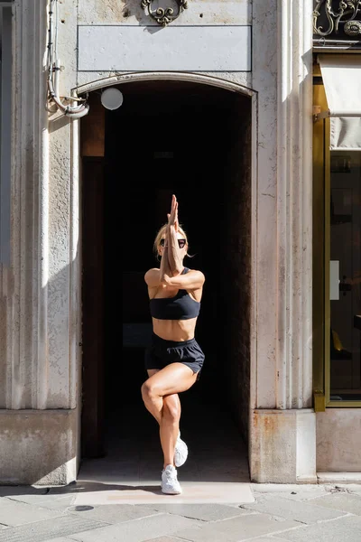 Deportista rubia en zapatillas blancas, gafas de sol, top negro y pantalones cortos haciendo yoga Eagle pose en la calle urbana de Venecia - foto de stock