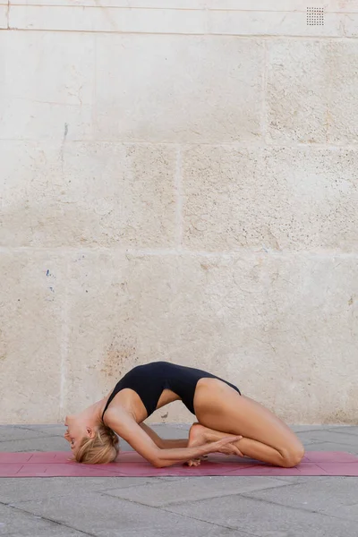 Vue latérale de la femme blonde pratiquant le yoga pose kapotasana sur le trottoir à Venise — Photo de stock