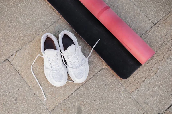 Vista superior de zapatillas blancas y alfombra de fitness rosa en la acera al aire libre - foto de stock