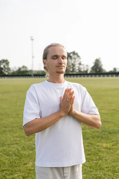 Giovane uomo in cotone bianco t-shirt meditando con gesto anjali mudra e gli occhi chiusi sullo stadio all'aperto — Foto stock