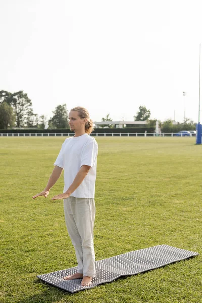 Homme aux cheveux longs en pantalon de lin et t-shirt blanc méditant les yeux fermés tout en se tenant debout sur le tapis de yoga sur la pelouse verte — Photo de stock