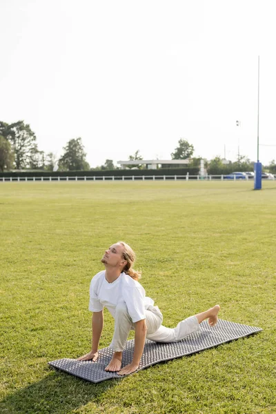 Longueur totale de l'homme en t-shirt blanc et pantalon de coton méditant dans la pose de fente avant sur tapis de yoga et herbe verte à l'extérieur — Photo de stock