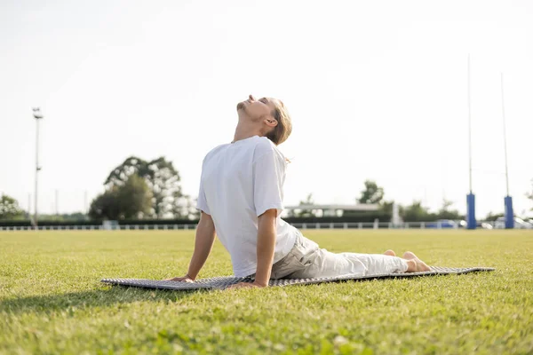 Nivel superficial del hombre joven del yoga que practica la pose de la cobra con los ojos cerrados en la hierba verde al aire libre - foto de stock