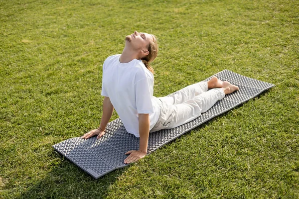 Full length of barefoot man in white t-shirt and linen pants practicing yoga in cobra pose on green lawn — Stock Photo