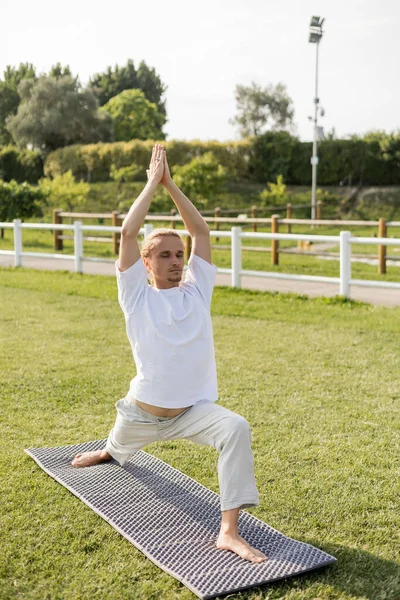 Longitud completa de hombre descalzo en pantalones de lino y camiseta blanca practicando yoga en pose guerrera con las manos levantadas sobre césped cubierto de hierba - foto de stock