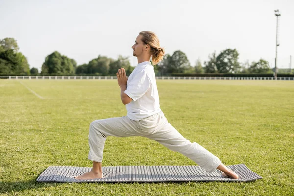 Vue latérale de l'homme pieds nus pratiquant le yoga en posture guerrière avec geste anjali mudra sur l'herbe verte du stade extérieur — Photo de stock