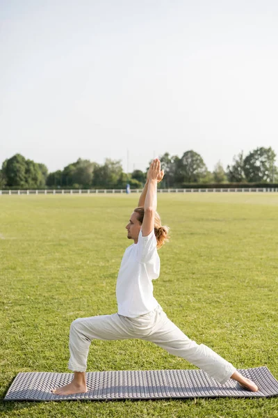 Vista lateral del hombre en pantalones de lino y camiseta blanca practicando pose guerrera con las manos orantes levantadas sobre estera de yoga en césped verde - foto de stock