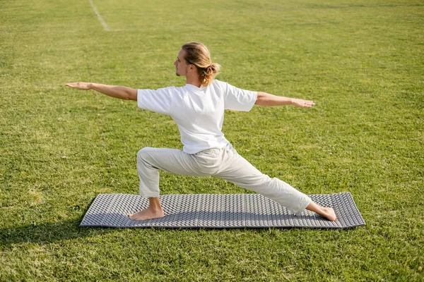 Side view of young long haired man in white t-shirt meditating in warrior pose with outstretched hands on yoga mat outdoors — Stock Photo