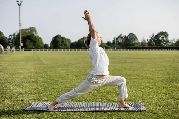 Side view of sportive barefoot man meditating in warrior pose with raised hands on stadium outdoors — Stock Photo