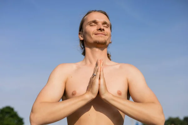 Low angle view of carefree shirtless man meditating with closed eyes and anjali mudra gesture against blue cloudless sky outdoors — Stock Photo