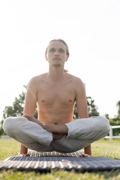 Молодой человек без рубашки в льняных брюках практикующий йогу в масштабе позы на коврике йоги на открытом воздухе — стоковое фото