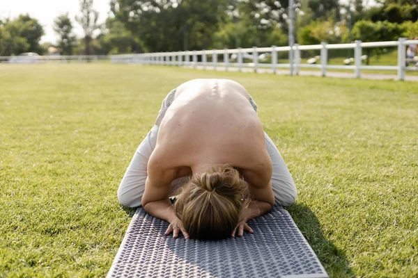 Shirtless man with long hair meditating in himalayan duck pose on yoga mat on grassy field — Stock Photo