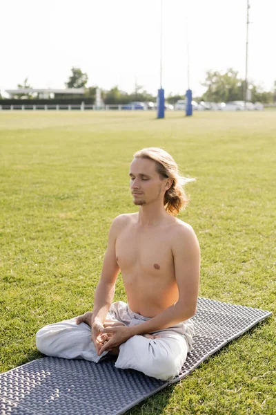 Hombre de pelo largo sin camisa meditando en pose de loto con los ojos cerrados mientras está sentado en la esterilla de yoga en el estadio cubierto de hierba - foto de stock