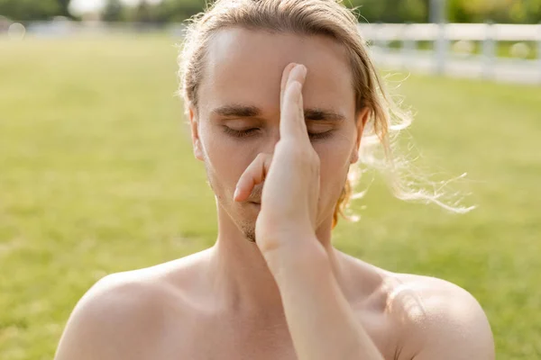 Young yoga man with bare shoulders and closed eyes holding hand near face and doing pranayama exercise outdoors — Stock Photo