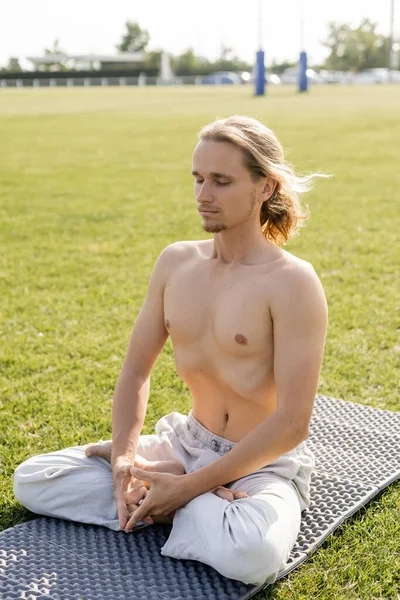 Hombre sin camisa de pelo largo en pantalones de lino meditando en pose de loto con los ojos cerrados en el estadio cubierto de hierba - foto de stock