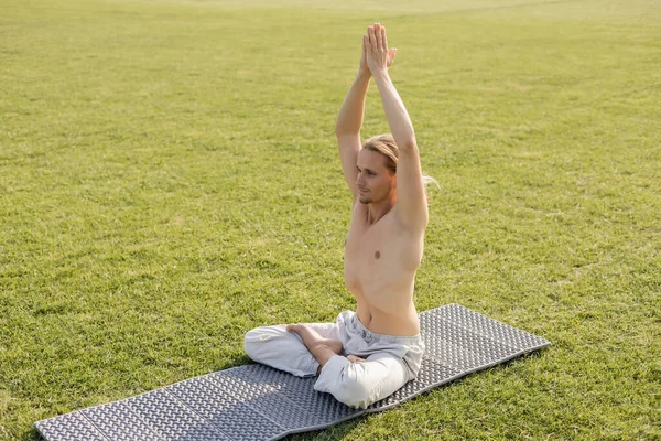 Счастливый мужчина без рубашки в хлопковых штанах сидит в позе лотоса с поднятыми руками, медитируя на коврике для йоги и зеленой траве на открытом воздухе — стоковое фото