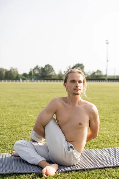 Hombre descalzo sin camisa en pantalones de lino mirando hacia otro lado mientras practica yoga en pose de salvia en el campo verde del estadio al aire libre - foto de stock